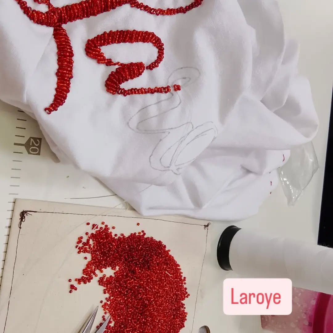 Bordado em miçanga vermelha na palavra Laroyé sobre tecido branco, com miçangas e pinça na mesa